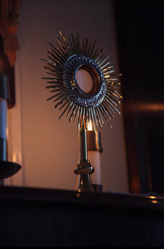 A picture of the Holy Eucharist in a gold and silver monstrance on an altar with a candle on the left and behind.