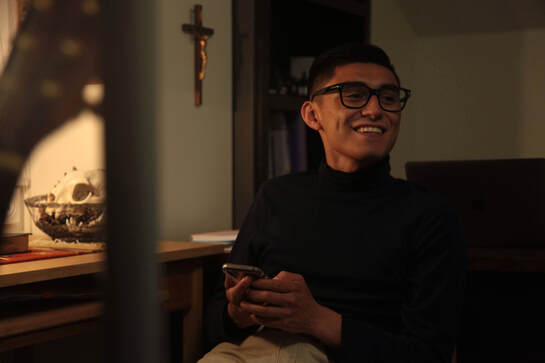 A picture of a young man on his phone, sitting and smiling wearing a dark turtleneck and glasses, with a dark book shelf and brown and gold crucifix on the wall behind him.