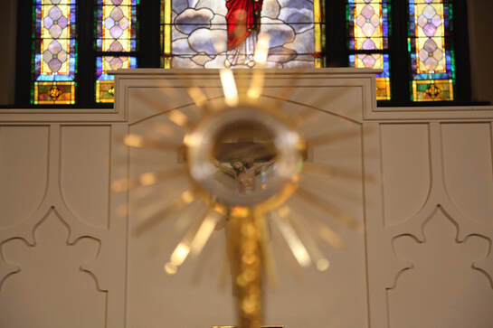 A picture with a crucifix torso-up, in-focus in the center, seen through an empty monstrance, with stained glass that is a rainbow of colors in the back.