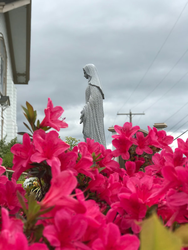 A picture of the left side of the grey statue of Mary, with grey cloudy skies, telephone wires on the right, the side of a church on the left and pink rhododendron flowers on the bottom half.
